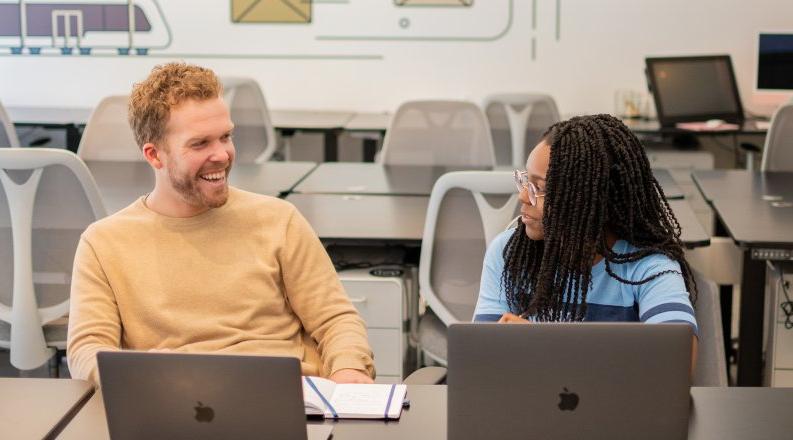 A female student laughs with a male supervisor in an office space while they look at laptops.