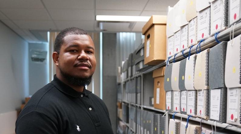 A student stands next to a bookshelf in a library.